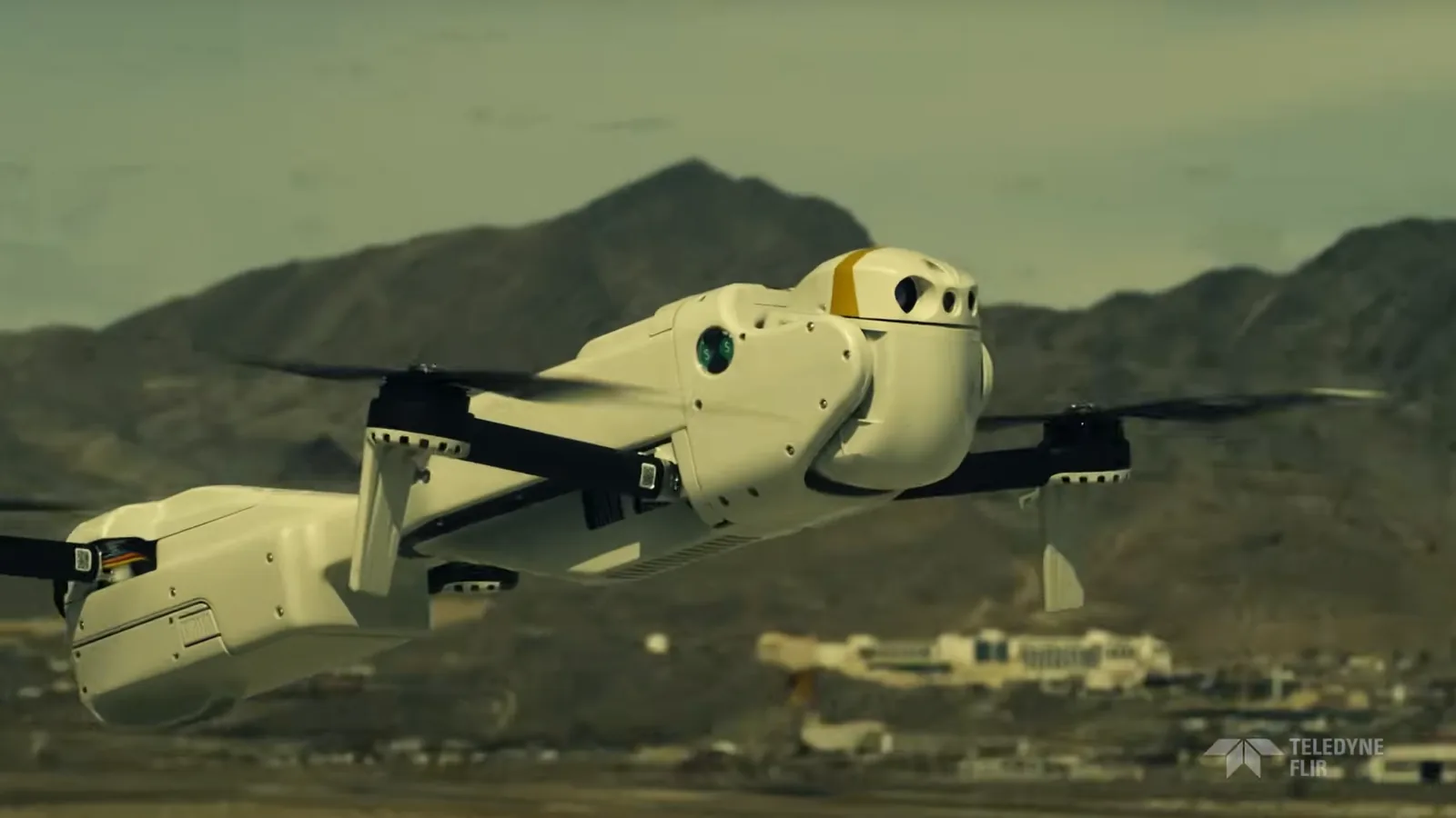 A white FLIR drone in flight low over the desert, with mountains behind